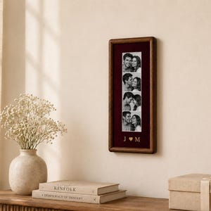 May include: A rectangular dark wood picture frame containing a vertical strip of four black and white photographs of a couple. Below the photos, the letters J and M are separated by a heart symbol in gold lettering on a deep burgundy background.