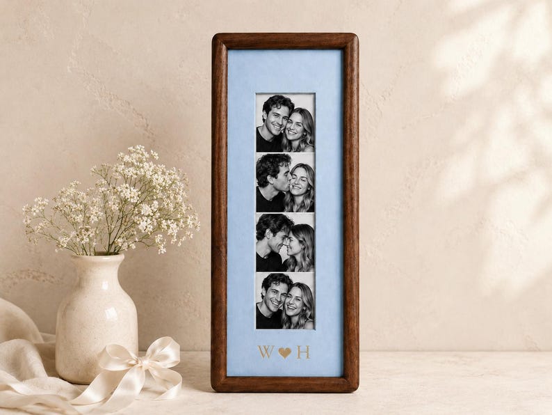May include: A vertical rectangular dark wood picture frame containing a light blue mat board with four stacked black and white photographs of a couple. The mat board features the gold-colored letters W and H separated by a heart symbol.