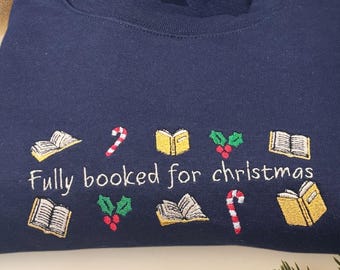 Puede incluir: Sudadera azul marino con bordados de libros, bastones de caramelo y acebo. El texto "Fully booked for christmas" está bordado debajo. La sudadera está doblada, rodeada de adornos navideños.