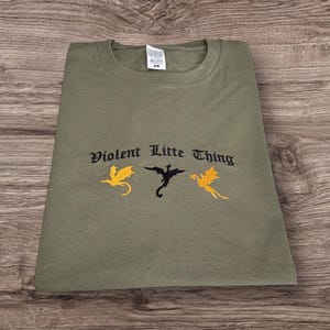 May include: An olive green t-shirt featuring the embroidered phrase "Violent Little Thing" and three dragon motifs in yellow and black. The shirt is neatly folded on a wooden surface.