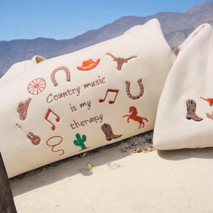 May include: Cream-coloured tote bags with country music embroidery. One bag has the text "Country music is my therapy" with musical notes, a horseshoe, a cowboy hat, and a cactus. The other bag has a cowboy boot and a horse.