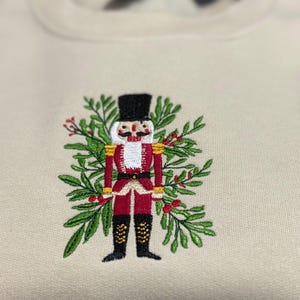 May include: Cream-coloured sweatshirt with an embroidered nutcracker soldier design. The nutcracker wears a red jacket, black boots, and a tall black hat, surrounded by green leaves and red berries. A festive holiday-themed garment.