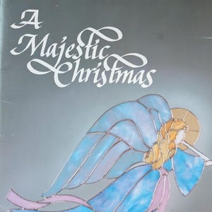 May include: A book cover with the title "A Majestic Christmas" in white script. It features a stained-glass style illustration of an angel in blue and gold, playing a trumpet. The author's name, Donna Schulze, is at the bottom.