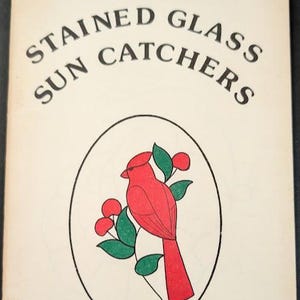 May include: A softcover book titled STAINED GLASS SUN CATCHERS VOLUME 1. The cover is off-white with black text and features a central oval illustration of a red cardinal bird perched on a green leafy branch with red berries.
