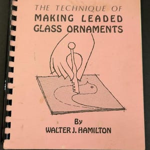 May include: A spiral-bound instructional book with a light pink cover titled The Technique of Making Leaded Glass Ornaments by Walter J. Hamilton. The cover features a black line drawing of a hand using a tool on a piece of glass.