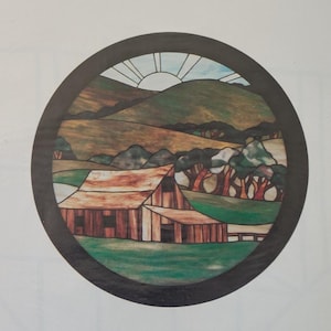 May include: A circular stained glass panel featuring a landscape scene with a brown barn in the foreground, rolling green and tan hills, a cluster of trees, and a sun rising over the horizon, all enclosed within a dark, solid circular frame.
