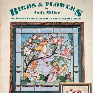 May include: A book cover titled Birds & Flowers by Judy Miller, 100 Designs for Stained Glass & Needle Arts. The cover features a stained glass panel with a bird perched on a branch surrounded by pink flowers, framed in a dark wood border.