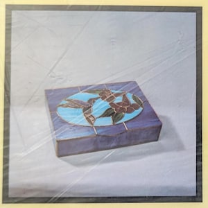 May include: A rectangular stained glass box featuring a central oval panel with a hummingbird and floral design in shades of blue, green, and brown. The box has deep purple glass sides and a hinged lid, constructed with visible metal solder lines.