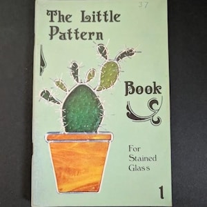 May include: A light green softcover book titled The Little Pattern Book For Stained Glass 1. The cover features a central illustration of a dark green cactus with white spines planted in an orange pot, with the number 37 printed at the top.