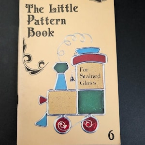 May include: A tan softcover book titled The Little Pattern Book For Stained Glass, featuring a graphic of a colorful steam train with red, blue, green, and yellow sections, black text, and the number 6 printed in the bottom right corner.