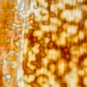May include: A close-up view of a translucent material featuring an organic, mottled pattern in shades of amber, burnt orange, cream, and soft brown. The surface displays a fluid, marbled texture with irregular, cloud-like shapes and light diffusion.