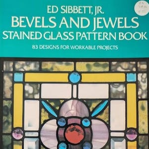 May include: A softcover book titled Bevels and Jewels Stained Glass Pattern Book by Ed Sibbett, Jr. The teal cover features white text and a graphic of a stained glass window with yellow, blue, pink, and clear glass pieces in a geometric design.