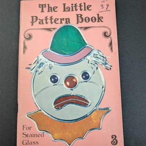 May include: A pink softcover book titled The Little Pattern Book For Stained Glass. The cover features a graphic illustration of a clown face with a green hat, blue eyes, a red nose, and an orange collar, with the number 39 written in ink.