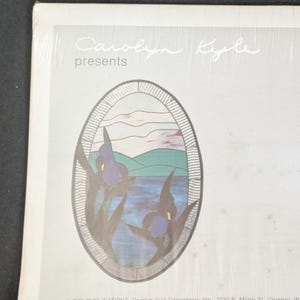 May include: A rectangular paper print featuring the text Carolyn Kyle presents at the top. Below the text is an oval illustration of a stained glass window depicting blue iris flowers, a body of water, and green hills under a pale sky.