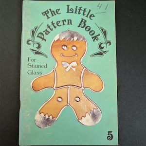 May include: A softcover book titled The Little Pattern Book For Stained Glass with a light green cover. The center features a brown gingerbread person illustration with a white bow tie and white accents on the hands and feet. The number 5 is at the bottom.