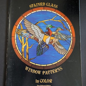 May include: A black book cover titled STAINED GLASS WINDOW PATTERNS in COLOR New Enlarged Edition. The center features a circular stained glass design of two ducks flying over blue water with yellow tree branches against a dark background.