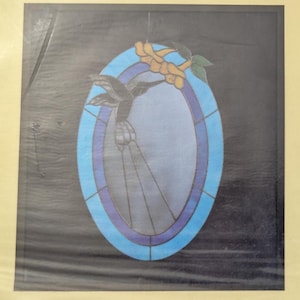 May include: An oval stained glass suncatcher featuring a dark hummingbird silhouette in flight. The piece has a light blue and purple border, with a yellow flower and green leaves at the top, all set against a dark, solid background.