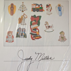 May include: A collection of Christmas-themed illustrations featuring angels, a tree, a train, a candle-bearing figure, bells with a red bow, a rocking horse, and dolls. The artwork is signed Judy Miller at the bottom in black cursive script.