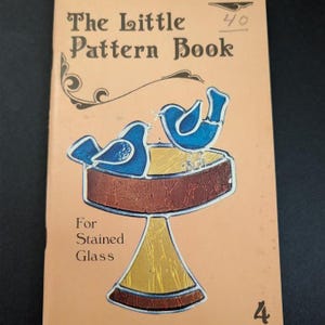 May include: A tan softcover book titled The Little Pattern Book For Stained Glass. The cover features a graphic illustration of two blue birds perched on a brown and yellow pedestal birdbath. The number 40 is written in the top right corner.