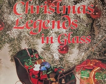 May include: A stained glass sleigh filled with miniature wrapped gifts and a small white bear figurine. The text reads "Carolyn Kyle presents Christmas Legends in Glass" against a background of a decorated Christmas tree with red ornaments.