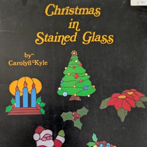 May include: A black book cover titled Christmas in Stained Glass by Carolyn Kyle. The cover features colorful stained glass illustrations of a Christmas tree, blue candles, a poinsettia, holly leaves, and a Santa Claus figure on a black background.