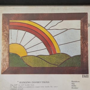 May include: A stained glass pattern featuring a yellow sun, a multi-colored rainbow with red and yellow bands, and green hills against a light blue sky. The design is enclosed in a brown frame and includes text for hanging instructions and labels.
