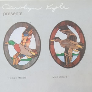 May include: Carolyn Kyle presents two oval stained glass panels featuring mallard ducks in flight. The left panel shows a female mallard, and the right panel shows a male mallard with a green head. Both designs are framed in dark wood.
