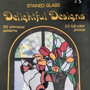 May include: A book cover titled Stained Glass Delightful Designs featuring 58 whimsical patterns and 24 full-color photos. The cover art displays a stained glass window illustration of a calico cat sitting next to a vase of red, white, and orange tulips.
