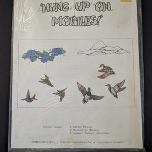 May include: A plastic-wrapped instructional pattern sheet titled Hung Up On Mobiles! featuring illustrations of stained glass hanging mobiles with blue flowers, hummingbirds, clouds, and ducks. Includes text listing contents and 1993 copyright info.
