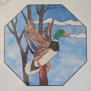 May include: An octagonal stained glass panel featuring a mallard duck in flight with brown, green, white, and orange glass pieces. The duck is positioned against a background of blue sky, white clouds, and dark brown tree branches with lead lines.