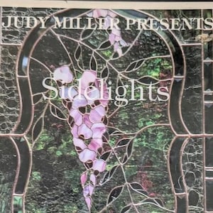 May include: A stained glass panel featuring a central cluster of light pink and purple wisteria flowers with green leaves, framed by dark, textured glass borders. The text JUDY MILLER PRESENTS and Sidelights is printed in white across the glass.