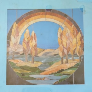 May include: A square stained glass panel featuring a circular landscape scene. The design depicts a blue sky, a brown arched rainbow, and tall yellow-brown trees flanking a central stream that flows through green and tan rolling hills.