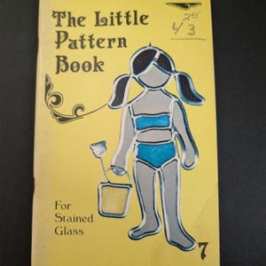 May include: A yellow softcover book titled The Little Pattern Book For Stained Glass. The cover features a graphic illustration of a person with pigtails wearing a blue two-piece swimsuit and holding a small bucket and shovel, with the number 7.