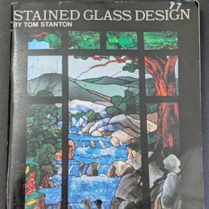 May include: A book titled STAINED GLASS DESIGN BY TOM STANTON featuring a cover illustration of a landscape with a flowing blue river, rocky banks, green trees, and a large orange-brown tree trunk, all framed by a black window pane design.