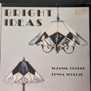 May include: A book cover titled BRIGHT IDEAS by Suzanne Cooper and Donna Schulze featuring illustrations of two stained glass lamps with geometric patterns in white and dark tones, suspended by a chain and standing on a base respectively.