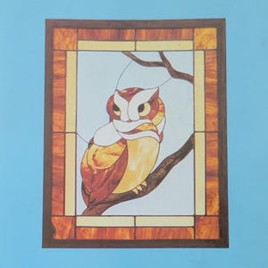 May include: A rectangular stained glass panel featuring an owl perched on a tree branch. The owl is rendered in shades of amber, brown, and white. The panel is framed by a border of mottled orange and brown glass with light yellow accents.