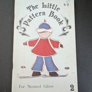 May include: A softcover book titled The Little Pattern Book For Stained Glass 2, featuring a light blue cover with a central illustration of a skier wearing a red jacket, blue pants, and a blue hat, holding ski poles on a pair of skis.