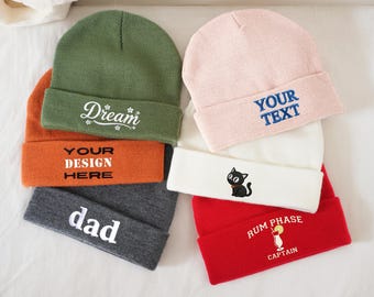 May include: A collection of colorful knit beanies. The beanies are in various colors, including green, orange, gray, pink, white, and red. Some beanies have text such as "Dream", "Your Design Here", "dad", and "Your Text". Other beanies have a cat graphic and a cocktail graphic.