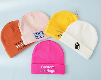May include: Five colorful beanies in orange, pink, yellow, white, and fuchsia. Each beanie has a different design, including text that says "YOU DESIGN HERE", "YOUR TEXT", "ALASKA", and a black cat graphic. The fuchsia beanie has the text "Custom text/logo".