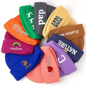 May include: A circular arrangement of eleven colorful knit beanies, each featuring unique embroidered text or graphics including 'Dream', 'dad', 'YOUR TEXT', 'Custom text', 'NATURE', 'C', 'LOVE' with a heart, and a lion logo.