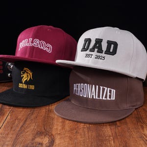 Könnte beinhalten: Drei Baseballkappen mit unterschiedlichen Designs. Die erste Kappe ist burgunderfarben mit weißem Text, der "CUSTOM" lautet. Die zweite Kappe ist weiß mit schwarzem Text, der "DAD EST 2025" lautet. Die dritte Kappe ist braun mit weißem Text, der "PERSONALIZED" lautet.