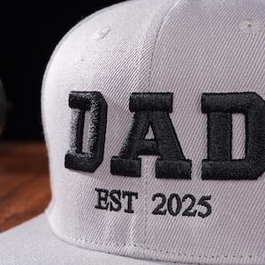 Może przedstawiać: Szara czapka bejsbol&oacute;wka z czarnym haftem z napisem "DAD EST 2025".
