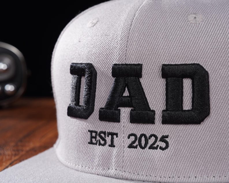 Może przedstawiać: Szara czapka bejsbol&oacute;wka z czarnym haftem z napisem "DAD EST 2025".