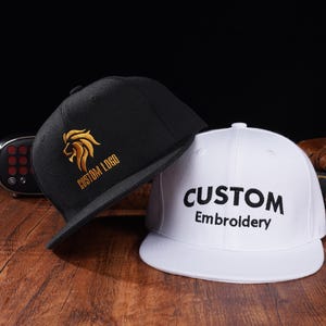Peut inclure: Deux casquettes de baseball noires et blanches avec du texte brodé. Une casquette a un logo de lion doré avec le texte "CUSTOM LOGO". L'autre casquette a le texte "CUSTOM Embroidery".