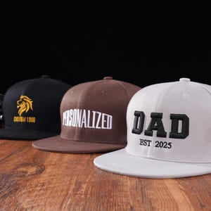Puede incluir: Tres gorras de béisbol con diseños diferentes. La primera gorra es negra con un logotipo de león amarillo y el texto "Custom Logo". La segunda gorra es marrón con texto blanco que dice "Personalized". La tercera gorra es blanca con texto negro que dice "DAD EST 2025".