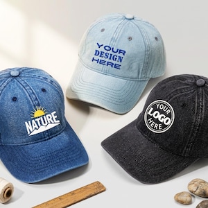 Könnte beinhalten: Drei Baseballkappen in verschiedenen Farben: Jeansblau, Hellblau und Schwarz. Die blaue Kappe hat ein "NATURE"-Logo, die hellblaue Kappe sagt "YOUR DESIGN HERE", und die schwarze Kappe hat ein "YOUR LOGO HERE"-Logo.