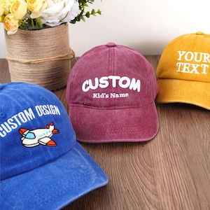 May include: Three baseball caps in blue, burgundy, and yellow. The blue cap has "CUSTOM DESIGN" and an airplane graphic. The burgundy cap says "CUSTOM Kid's Name". The yellow cap says "YOUR TEXT".