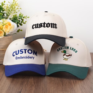 May include: Three baseball caps with custom embroidery. The caps have a cream-colored crown and a colored brim. One cap has a black brim and the word "custom" embroidered in black. Another has a blue brim with "CUSTOM Embroidery" in blue. The third has a green brim with a duck logo.
