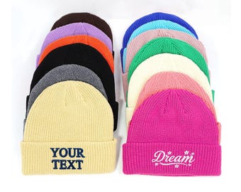 May include: A collection of colorful knit beanies in various shades, including black, gray, and pink. One beanie is embroidered with "YOUR TEXT" in navy blue, while another features "Dream" in white script with floral accents. The beanies are stacked.