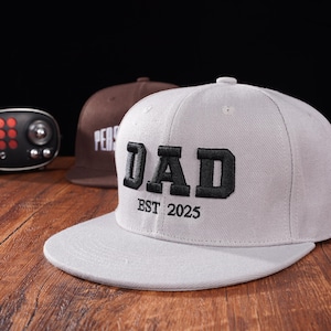 Könnte beinhalten: Eine graue Baseballkappe mit einem schwarzen gestickten "DAD" und "EST 2025" auf der Vorderseite.